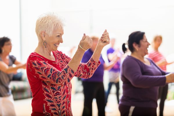 Beautiful senior woman snaps her fingers as she learns new dance moves during dance class at her senior center. People are dancing in the background.